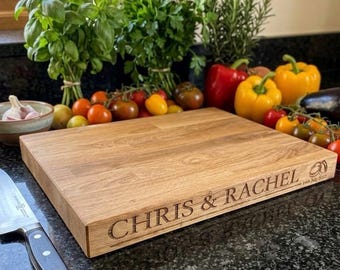 Puede incluir: Una tabla de cortar rectangular de madera con veta marrón claro, que presenta el texto grabado "CHRIS & RACHEL" y un pequeño icono de anillos con la fecha "14th July 2023" en el borde frontal, situada junto a un cuchillo de cocina.