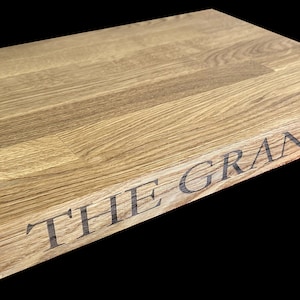 Op de afbeelding: Een rechthoekige houten snijplank met de tekst "THE GRANARY" gegraveerd op de rand. De plank heeft een lichte bruine kleur en een natuurlijke houtnerf. De oppervlakte is glad.