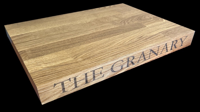 Op de afbeelding: Een rechthoekige houten snijplank met de tekst "THE GRANARY" gegraveerd op de rand. De plank heeft een lichte bruine kleur en een natuurlijke houtnerf. De oppervlakte is glad.