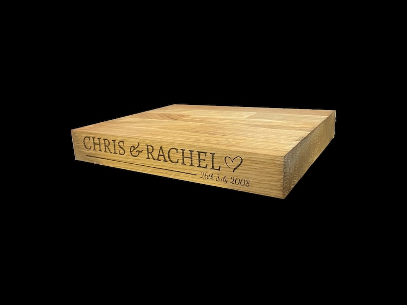 Op de afbeelding: Een lichtgekleurd houten blok met gegraveerde tekst. De tekst luidt "CHRIS & RACHEL" boven een hartsymbool en de datum "26th July 2008". Het blok is rechthoekig en lijkt een decoratief item te zijn.