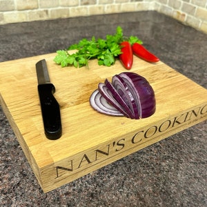 Op de afbeelding: Een rechthoekige houten snijplank met de gegraveerde tekst "NAN'S COOKING is the best". Het heeft een zwart mes, gesneden rode ui, rode chilipepers en verse groene peterselie. De plank is van lichtgekleurd hout.