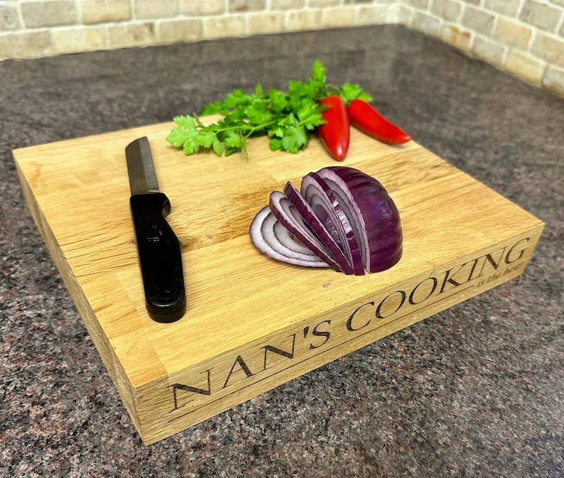 Op de afbeelding: Een rechthoekige houten snijplank met de gegraveerde tekst "NAN'S COOKING is the best". Het heeft een zwart mes, gesneden rode ui, rode chilipepers en verse groene peterselie. De plank is van lichtgekleurd hout.