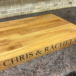 Op de afbeelding: Een rechthoekige houten snijplank met gegraveerde tekst "CHRIS & RACHEL" en de datum "26th July 2008". Het bord heeft ook een gegraveerd ontwerp van twee trouwringen. Het hout heeft een natuurlijke, lichtbruine kleur.