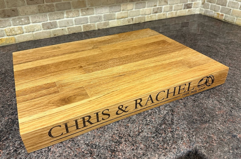 Op de afbeelding: Een rechthoekige houten snijplank met gegraveerde tekst "CHRIS & RACHEL" en de datum "26th July 2008". Het bord heeft ook een gegraveerd ontwerp van twee trouwringen. Het hout heeft een natuurlijke, lichtbruine kleur.