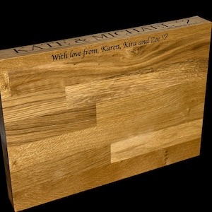 Op de afbeelding: Een lichtbruin houten blok met een natuurlijke houtnerf. De bovenkant is gegraveerd met de namen "KATE & MICHAEL" en een bericht, "With love from, Karen, Kira and Zoe" met een hartsymbool.