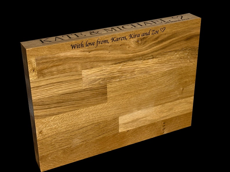 Op de afbeelding: Een lichtbruin houten blok met een natuurlijke houtnerf. De bovenkant is gegraveerd met de namen "KATE & MICHAEL" en een bericht, "With love from, Karen, Kira and Zoe" met een hartsymbool.