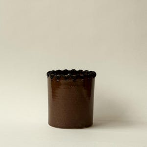 Op de afbeelding: Een cilindervormige bruine keramische vaas met een decoratieve zwarte rand rond de bovenkant. De vaas heeft een glanzende afwerking en staat op een neutrale achtergrond. Deze handgemaakte vaas is geschikt voor woondecoratie.