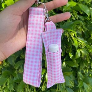 May include: Pink and white gingham keychain with a small pocket for a lip balm. The keychain has a silver metal ring and a silver metal clasp.