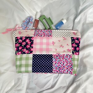 May include: A patchwork cosmetic bag with a variety of patterns, including floral, gingham, and polka dots. The bag contains beauty products like lip gloss, a hair clip, and a scrunchie. A pink bow is attached to the zipper.
