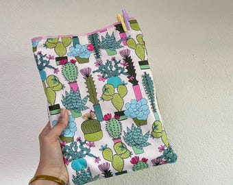 May include: A white fabric pouch with a colorful cactus print. The pouch features various cacti in shades of green, blue, and pink. A pink trim lines the top edge, and two pens are visible. The pouch is held by a person.