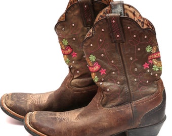 Può includere: Stivali da cowboy in pelle marrone con ricami rosa, verdi e gialli. Gli stivali hanno un design a stella e ferro di cavallo con strass.