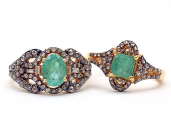 May include: Two antique-style rings with emerald center stones. One ring features an oval emerald, the other a square emerald. Both rings have a gold band and are surrounded by small, sparkling stones.