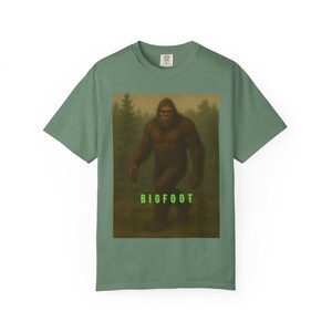 May include: Sage green t-shirt with a Bigfoot graphic. The design features a brown, hairy Bigfoot in a forest scene, with the word "BIGFOOT" in bright green below. The shirt has a crew neck and short sleeves, typical of a casual tee.