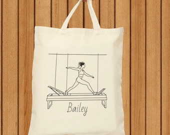 Può includere: Una borsa tote in tela bianca con un disegno a linea nera di una persona che fa pilates su un reformer. La borsa ha il nome "Bailey" stampato su di essa.