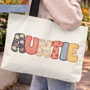 May include: A white tote bag with the word "AUNTIE" in colorful, patchwork-style letters. The letters feature floral, gingham, bow, strawberry, striped, and checkered patterns. A matching cosmetic bag is also available.