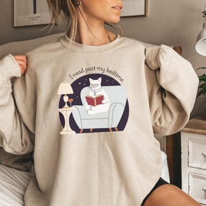 May include: A beige sweatshirt with a graphic of a cat reading a book in a chair. The text on the sweatshirt says "I read past my bedtime."