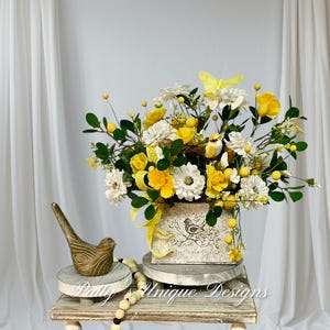 May include: A floral arrangement featuring yellow and white flowers, green leaves, and a yellow butterfly. The arrangement is in a decorative box with a bird illustration. A wooden bird figurine and a beaded garland are also present.