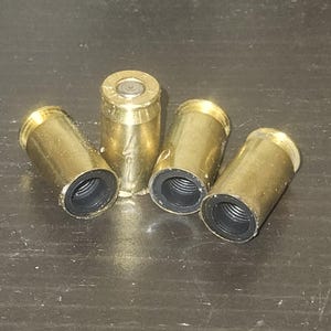 May include: Four brass bullet casings with black rubber inserts. The casings are arranged on a dark surface. The casings are cylindrical with a slightly flared rim and a small indentation on the top.