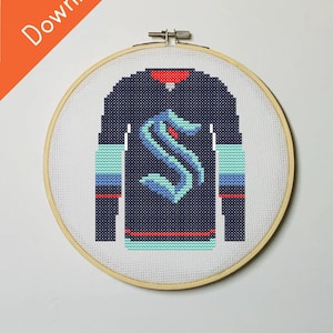 Op de afbeelding: Kruissteekpatroon van een marineblauw hockey-shirt met turquoise en rode accenten, met het Seattle Kraken-logo. Het ontwerp wordt gepresenteerd in een houten borduurring. De tekst "PDF Download" is zichtbaar.