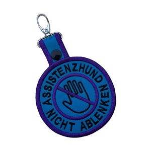 Könnte beinhalten: Ein blau-violetter Schlüsselanhänger mit dem Text "ASSISTENZHUND NICHT ABLENKEN" in einem kreisförmigen Design. Das Design enthält eine Hand mit einem Strich, der "Nicht ablenken" anzeigt. Der Schlüsselanhänger hat einen silbernen Verschluss.