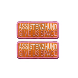 Könnte beinhalten: Zwei gestickte Aufnäher mit dem Text "ASSISTENZHUND GIVE US SPACE". Die Aufnäher haben einen orangefarbenen Hintergrund mit weißen Buchstaben und einen rosa Rand. Die Aufnäher sind rechteckig.