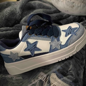 Könnte beinhalten: Weiße und blaue Sneaker mit Sternen. Die Schuhe haben eine weiße Basis mit Jeans-ähnlichen blauen Sternapplikationen und -besätzen. Dunkelblaue Schnürsenkel und eine weiße Sohle vervollständigen das Design. Die Sneaker sind neu.