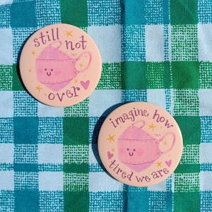 May include: Two pink and white button pins with a cartoon face mask design. The first pin says "still not over" and the second pin says "imagine how tired we are".