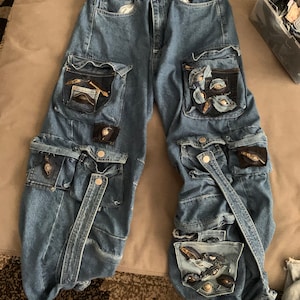May include: Blue denim cargo pants with multiple pockets and decorative embellishments. The pockets have zippers and flaps. The embellishments include gold-colored metal pieces and fabric patches.