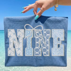 May include: A blue t-shirt with the word "NINE" in white and light blue, decorated with starfish, seashells, and a bow. The shirt is held up against a beach background. The text "Blue Jean" is at the bottom right.
