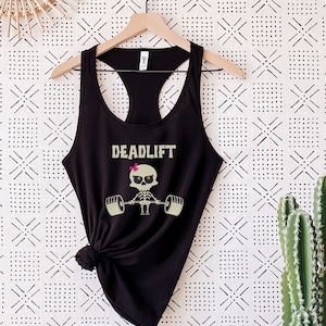 May include: Black racerback tank top with the word "DEADLIFT" in a tan font. A cartoon skeleton with a pink bow is lifting weights. The tank top is hanging on a wooden hanger against a patterned white and black background.