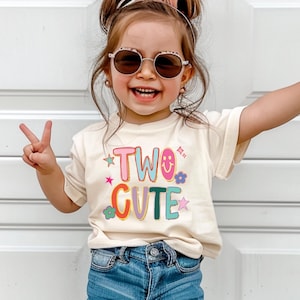 May include: A young child wearing a cream-colored t-shirt with the words "TWO CUTE" in colorful letters and graphics. The child is wearing sunglasses and blue denim shorts, making a peace sign with both hands. The background is a white door.