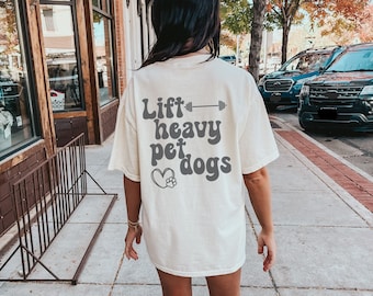 May include: Cream-colored t-shirt with the text "Lift heavy pet dogs" in gray. The design includes a barbell and a paw print. The shirt is a casual, oversized fit.
