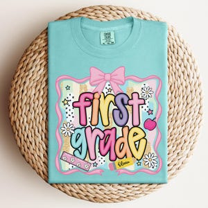 Op de afbeelding: Een lichtblauw t-shirt met de woorden "first grade" in kleurrijke bubbelletters. Het ontwerp bevat een roze strik, appel, madeliefjes, sterren en een liniaal. Het shirt ligt op een geweven, cirkelvormig oppervlak.