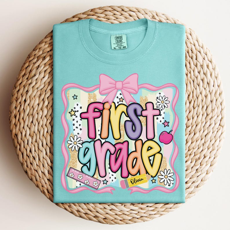 Op de afbeelding: Een lichtblauw t-shirt met de woorden "first grade" in kleurrijke bubbelletters. Het ontwerp bevat een roze strik, appel, madeliefjes, sterren en een liniaal. Het shirt ligt op een geweven, cirkelvormig oppervlak.