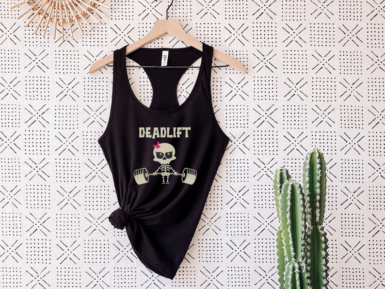 May include: Black racerback tank top with the word "DEADLIFT" in a tan font. A cartoon skeleton with a pink bow is lifting weights. The tank top is hanging on a wooden hanger against a patterned white and black background.