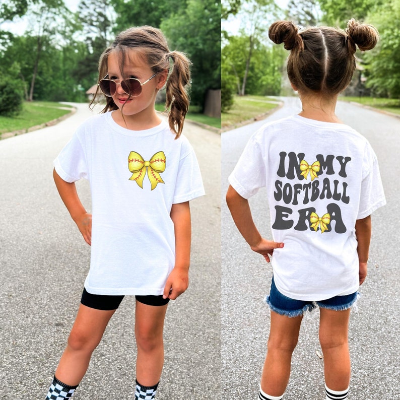 K&ouml;nnte beinhalten: Wei&szlig;es T-Shirt mit einer gelben Softball-Schleife auf der Vorderseite und dem Text "In My Softball Era" mit einer gelben Softball-Schleife auf der R&uuml;ckseite.