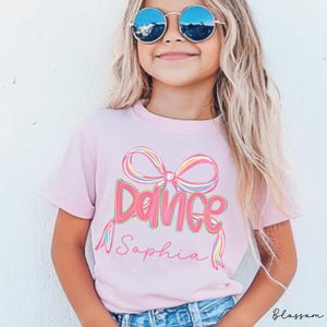 May include: A light pink t-shirt with the word "Dance" in a colorful, outlined font, and a matching bow graphic. The name "Sophia" is written below in a cursive font. The shirt is worn by a young person wearing sunglasses.