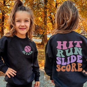 May include: Black sweatshirt with a baseball graphic and the name "Skylar" in teal. The back of the sweatshirt has the words "Hit Run Slide Score" in pink, teal, purple, and orange. The sweatshirt is worn by a young person.