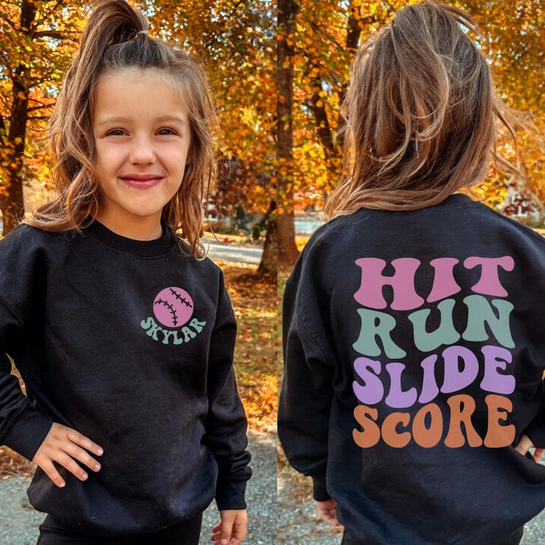 May include: Black sweatshirt with a baseball graphic and the name "Skylar" in teal. The back of the sweatshirt has the words "Hit Run Slide Score" in pink, teal, purple, and orange. The sweatshirt is worn by a young person.