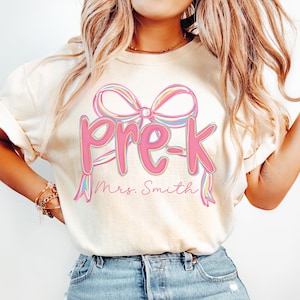 May include: Cream-colored t-shirt featuring the word "Pre-K" in pink script with a bow design above it. The name "Mrs. Smith" is written below the text in a cursive font. The shirt is a casual style.