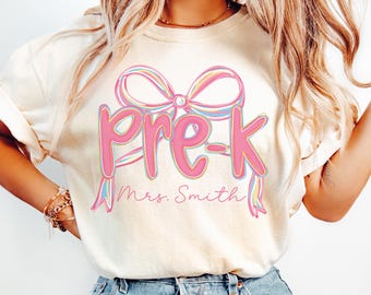 May include: Cream-colored t-shirt featuring the word "Pre-K" in pink script with a bow design above it. The name "Mrs. Smith" is written below the text in a cursive font. The shirt is a casual style.