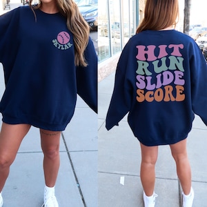 May include: Navy blue sweatshirt with a baseball graphic and the name "Skylar" on the front. The back of the sweatshirt features the words "Hit Run Slide Score" in a retro font. The sweatshirt is a comfortable, oversized fit.