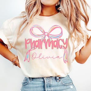 May include: Cream-colored t-shirt featuring the word "Pharmacy" in pink script with a rainbow bow design above it. The name "Olivia" is written below in a cursive font. The shirt is made of a soft material.