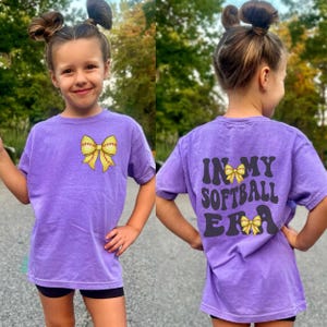 K&ouml;nnte beinhalten: Lilafarbenes Softball-T-Shirt mit einer gelben Softball-Schleife auf der Vorderseite und dem Text "In My Softball Era" mit einer gelben Softball-Schleife auf der R&uuml;ckseite.