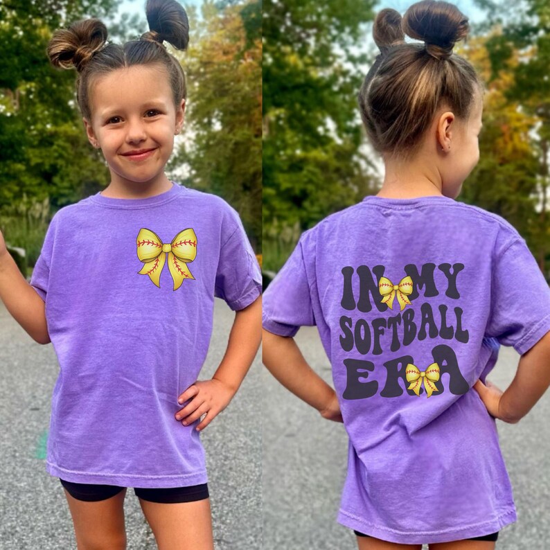 K&ouml;nnte beinhalten: Lilafarbenes Softball-T-Shirt mit einer gelben Softball-Schleife auf der Vorderseite und dem Text "In My Softball Era" mit einer gelben Softball-Schleife auf der R&uuml;ckseite.