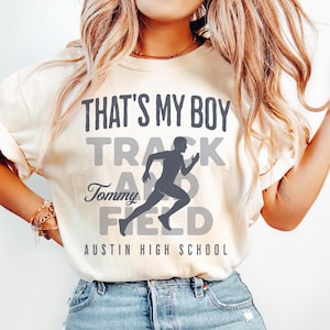 May include: Cream-colored t-shirt with the text "THAT'S MY BOY TRACK AND FIELD" in gray. A running figure graphic is in the center. The text "Tommy" and "Austin High School" are also included. The shirt is worn with blue jeans.
