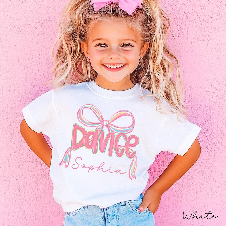 May include: White t-shirt with the word "Dance" in pink script and a colorful bow graphic. The name "Sophia" is written below the word "Dance". The shirt is worn by a young girl with blonde hair and a pink bow.