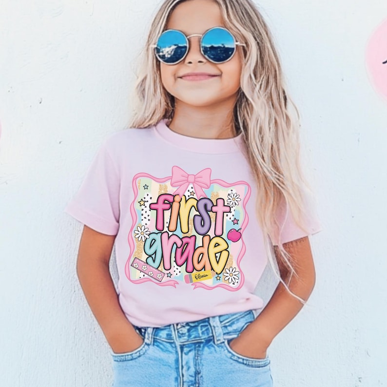 Op de afbeelding: Een lichtroze t-shirt met de woorden "first grade" in kleurrijke letters. Het ontwerp bevat een roze strik, bloemen, harten en potloden. Het shirt wordt gedragen door een jong persoon die een spijkerbroek en een zonnebril draagt.