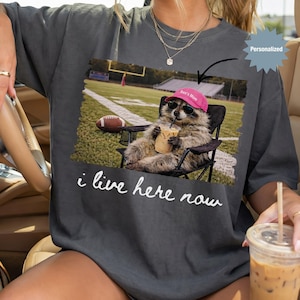Op de afbeelding: Donkergrijs t-shirt met een afbeelding van een wasbeer met een zonnebril en een roze hoed, ontspannen in een stoel met een drankje, op een voetbalveld. De tekst luidt "i live here now".