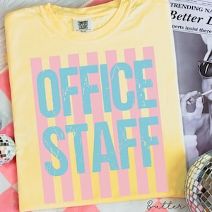 May include: A yellow Comfort Colors t-shirt with the words "OFFICE STAFF" in distressed blue letters over pink vertical stripes. The shirt is folded and lies on a striped surface.